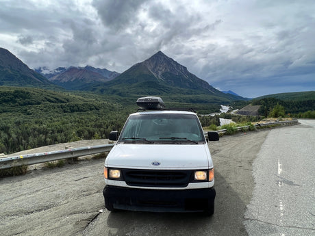 Scenic view with an rv rental: a white Ford van parked roadside against a backdrop of mountains, forests, and a river.