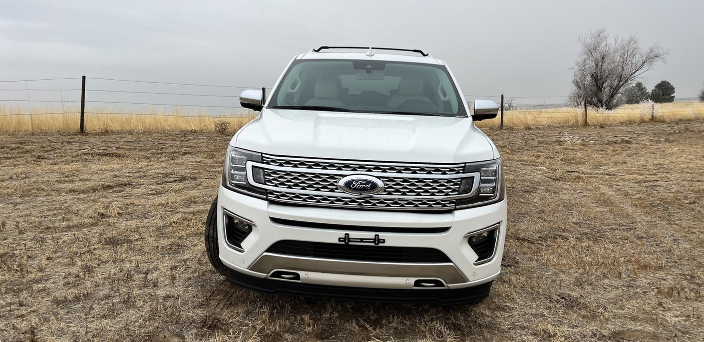 A white Ford Expedition SUV sits in a dry field under a cloudy sky. A spacious vehicle for trips, like an RV rental adventure.