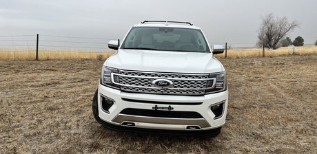 A white Ford Expedition SUV sits in a dry field under a cloudy sky. A spacious vehicle for trips, like an RV rental adventure.