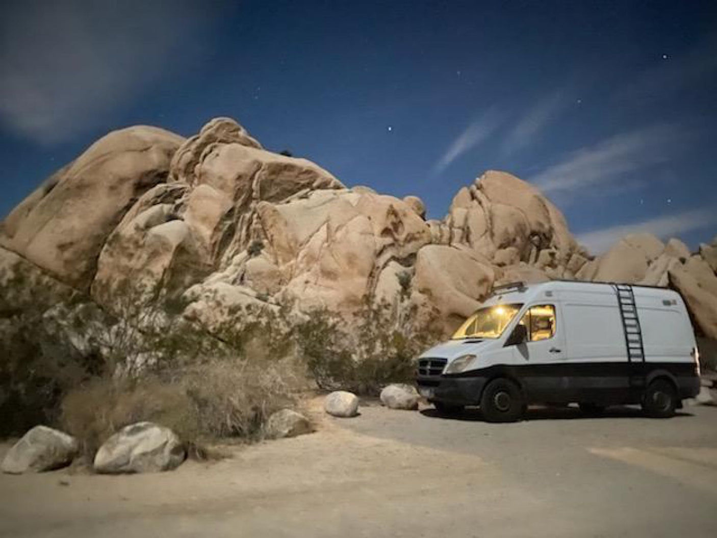 An RV rental camper van is parked at night among huge desert rock formations, with interior lights on and stars visible overhead.