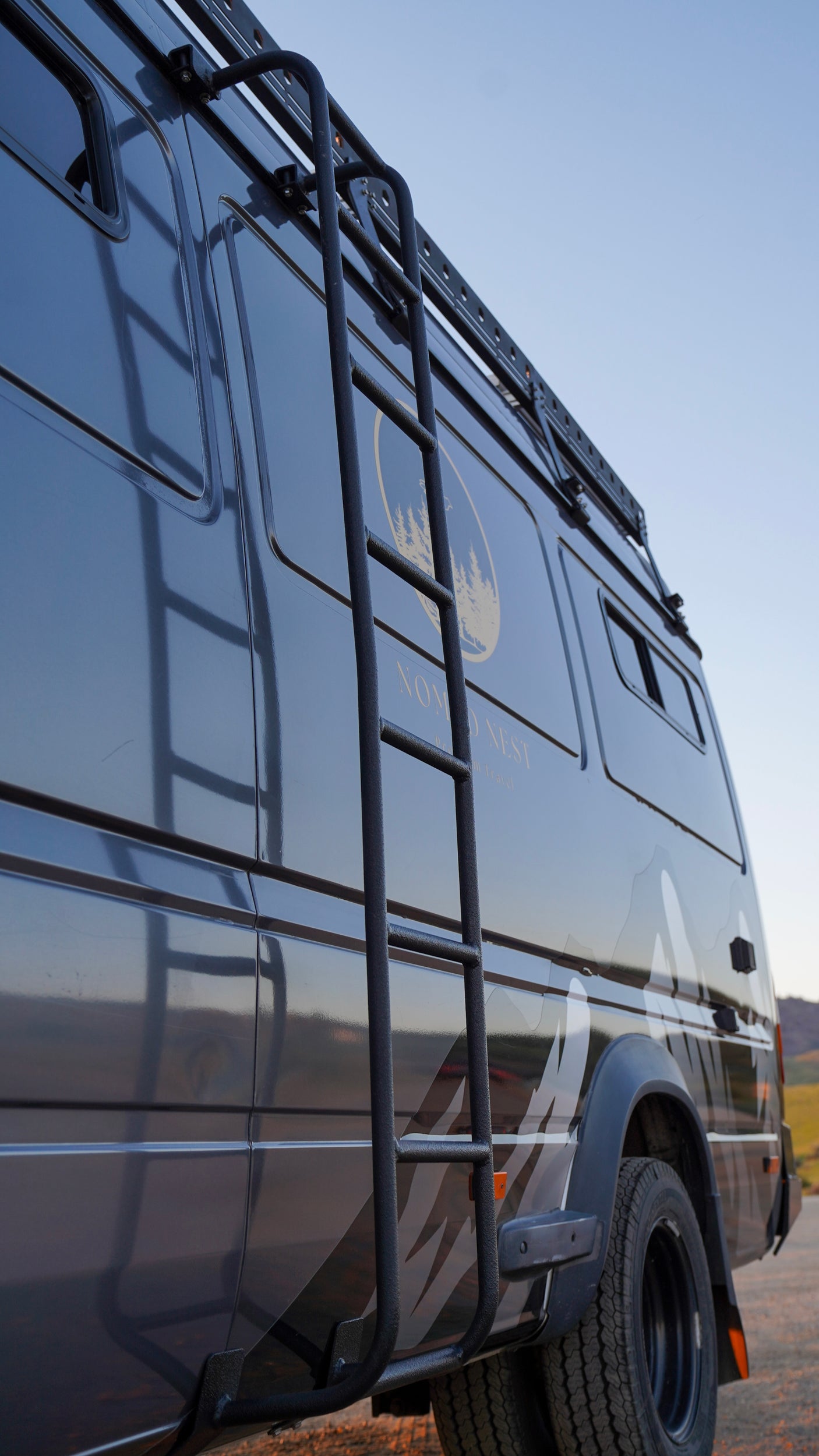 Low angle of a dark grey RV rental camper van with a side ladder, roof rack, and a unique 'Nomad Nest' logo. Great for travel!