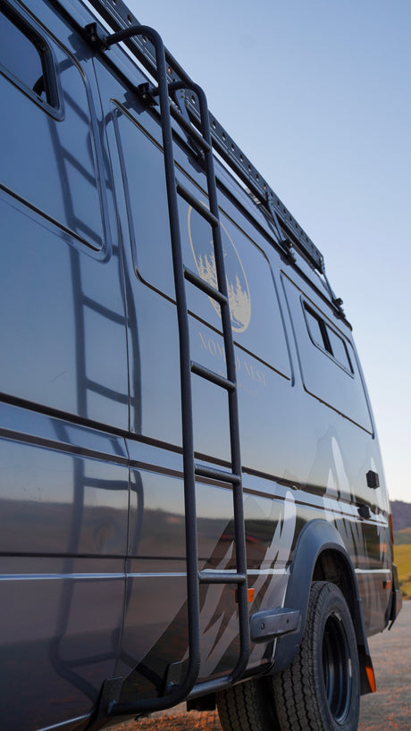 Low angle of a dark grey RV rental camper van with a side ladder, roof rack, and a unique 'Nomad Nest' logo. Great for travel!
