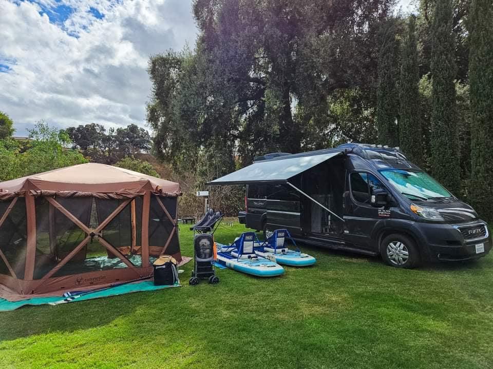  side door open to reveal the interior kitchenette. Ideal for|RV rental set up with brown screen tent & blue paddleboards on green grass — photo 2