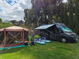 RV rental set up with brown screen tent & blue paddleboards on green grass, under cloudy skies.