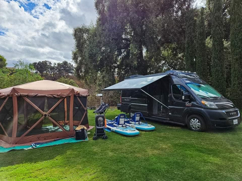  side door open to reveal the interior kitchenette. Ideal for|RV rental set up with brown screen tent & blue paddleboards on green grass — photo 2