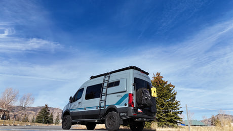 Adventure-ready gray Mode 4x4 RV rental van with off-road tires, ladder, and gear, parked under a clear blue sky.