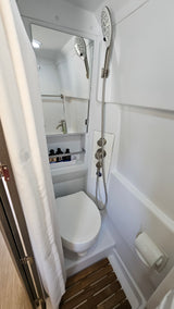 Compact RV rental wet bath with toilet, handheld shower, and sink (reflected in mirror). Features shower curtain & controls.
