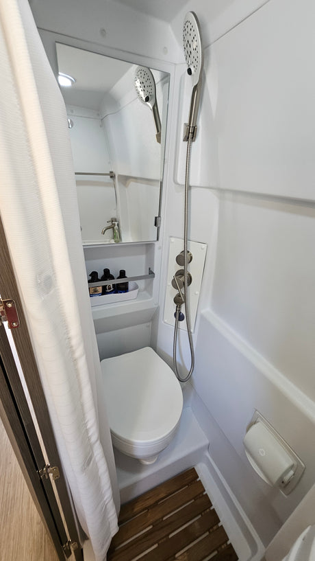 Compact RV rental wet bath with toilet, handheld shower, and sink (reflected in mirror). Features shower curtain & controls.