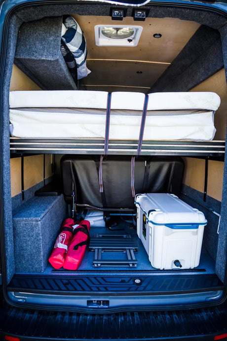 Rear view of an RV rental: features a high bed platform with stacked mattresses, a seat, cooler, & gear.