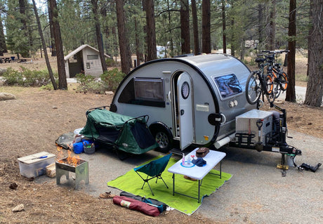 An RV rental teardrop camper campsite, complete with a fire pit, camping gear, and bikes, set among pine trees.