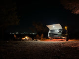 Cozy night camping with an RV rental: a bright travel trailer with open kitchen by a crackling campfire and chairs.