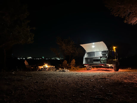 Cozy night camping with an RV rental: a bright travel trailer with open kitchen by a crackling campfire and chairs.