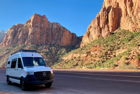 White RV rental van parked on a scenic road, surrounded by majestic red rock mountains under a blue sky.