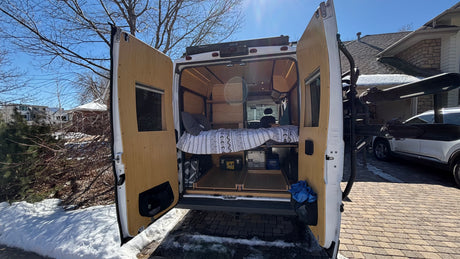 This RV rental camper van shows its cozy bed and storage conversion from the rear, parked on a snowy day.
