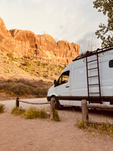 White camper van RV rental parked on a dirt road, facing towering desert rock formations.