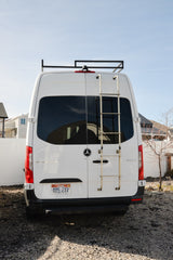 White Mercedes Sprinter campervan (RV rental) rear view, with roof rack & ladder, parked on gravel. Utah license plate.