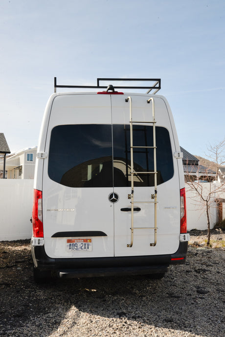 White Mercedes Sprinter campervan (RV rental) rear view, with roof rack & ladder, parked on gravel. Utah license plate.
