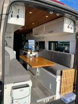 Stylish **RV rental** for van life: Features a wooden dinette, gray benches, kitchenette, and a scenic harbor view.