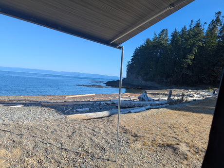 Enjoying a peaceful RV rental view of a sunlit beach, calm ocean, and forested coastline from under the awning.