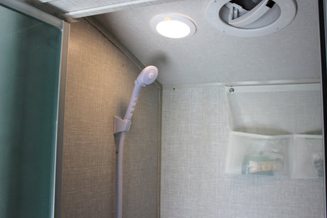 Compact RV rental shower interior: handheld shower head, ceiling light, ventilation fan, and wall-mounted mesh storage.