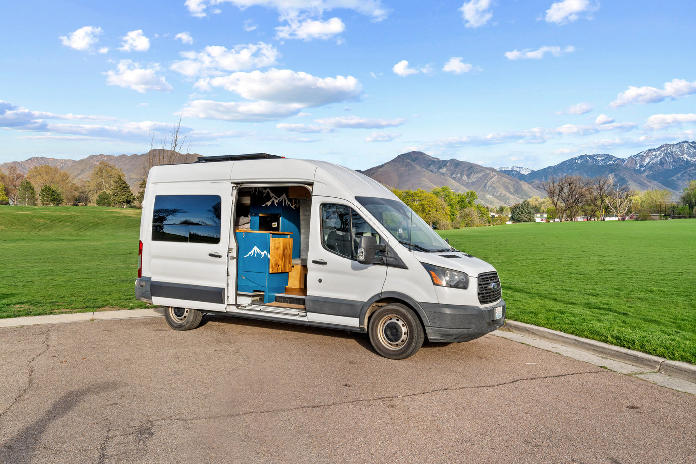 White Ford Transit camper van RV rental, with custom blue mountain-themed interior visible, parked by a green field with mountains in the background.