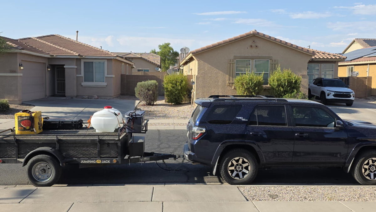 Dark blue SUV with utility trailer (water tank, gear) in a sunny neighborhood. No RV shown, but useful for rv rental trips.