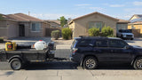Dark blue SUV with utility trailer (water tank, gear) in a sunny neighborhood. No RV shown, but useful for rv rental trips.