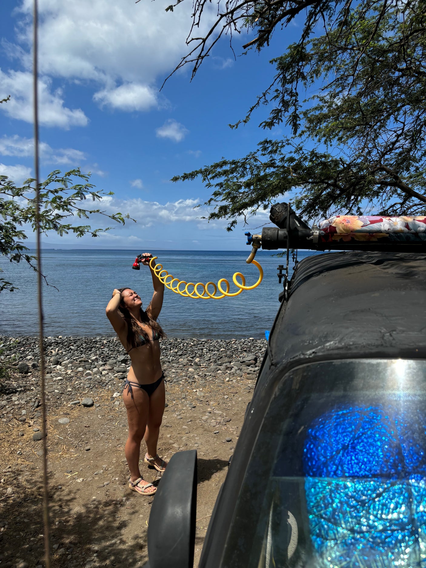 RV rental outdoor shower on a sunny beach. A woman in a bikini uses a yellow coiled hose by the sea.