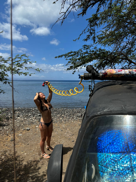 RV rental outdoor shower on a sunny beach. A woman in a bikini uses a yellow coiled hose by the sea.