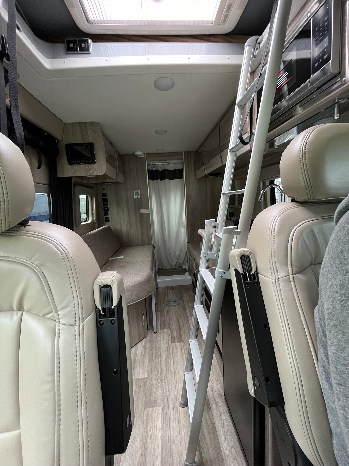 An rv rental's interior, featuring light-colored front seats, a ladder to an upper bunk, kitchenette with microwave, and a rear shower.