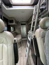 An rv rental's interior, featuring light-colored front seats, a ladder to an upper bunk, kitchenette with microwave, and a rear shower.