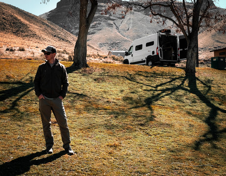 Scenic rv rental adventure. Man stands near his white camper van with open rear doors, parked in a grassy, mountainous landscape.