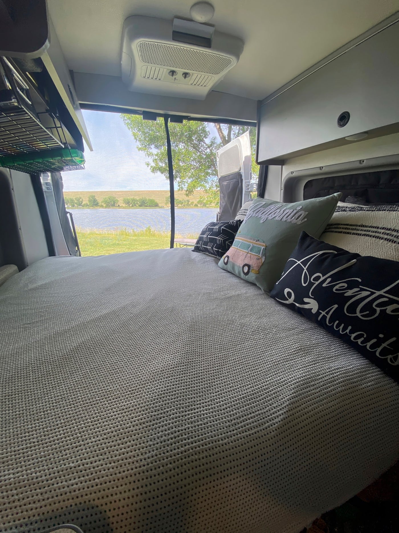 Inside an RV rental: a comfortable bed with travel-themed pillows overlooks a serene lake and green landscape. — photo 11