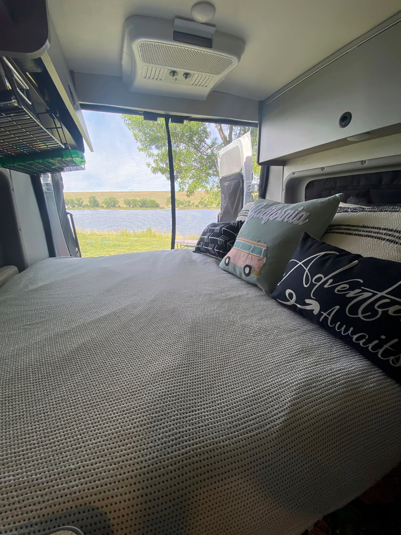 Inside an RV rental: a comfortable bed with travel-themed pillows overlooks a serene lake and green landscape.