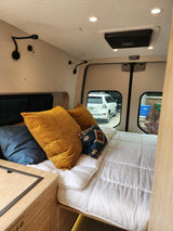 RV rental interior: A comfy bed with gold pillows, wood cabinetry, and a view out the back windows to the outside.