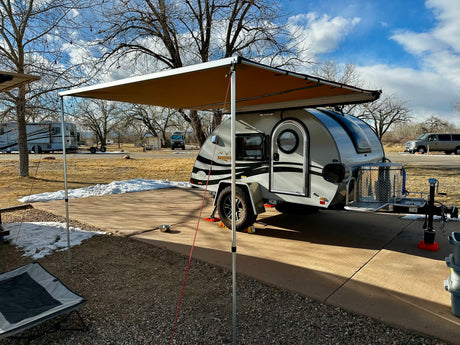 This RV rental teardrop trailer, with an extended awning, is parked at a snowy campsite under a blue sky, ready for adventure.