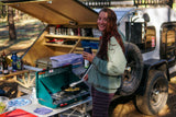 Happy woman cooks by her RV rental teardrop camper, enjoying the outdoor kitchen setup.