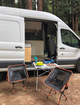 White camper van RV rental set up for outdoor cooking & dining. Portable stove, table, and chairs in a serene forest setting.