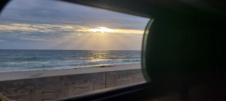 Scenic ocean view from an RV rental window, showing sunlight on the water, beach, and a patterned concrete barrier.