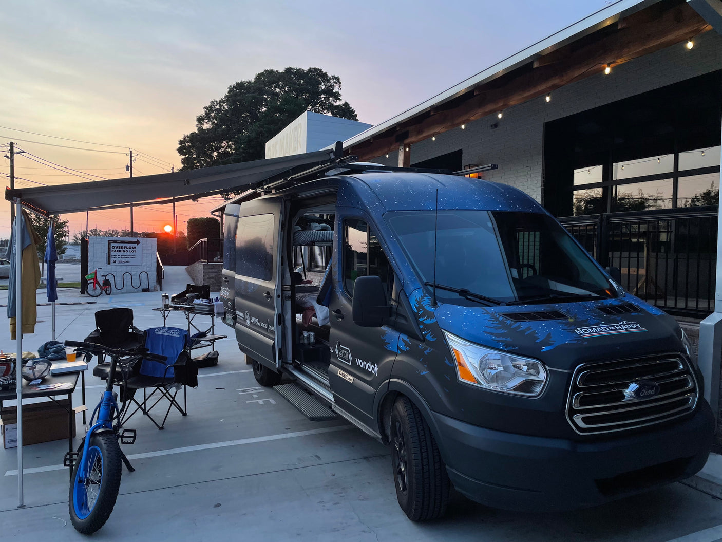 A custom dark blue Ford Transit rv rental van with an extended awning and outdoor gear, including a bike, is set up at sunset.
