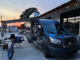 A custom dark blue Ford Transit rv rental van with an extended awning and outdoor gear, including a bike, is set up at sunset.