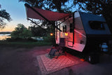 Relaxing at a lakefront RV rental campsite at dusk. An RV with an illuminated awning, chairs, and a rug overlook a calm lake.