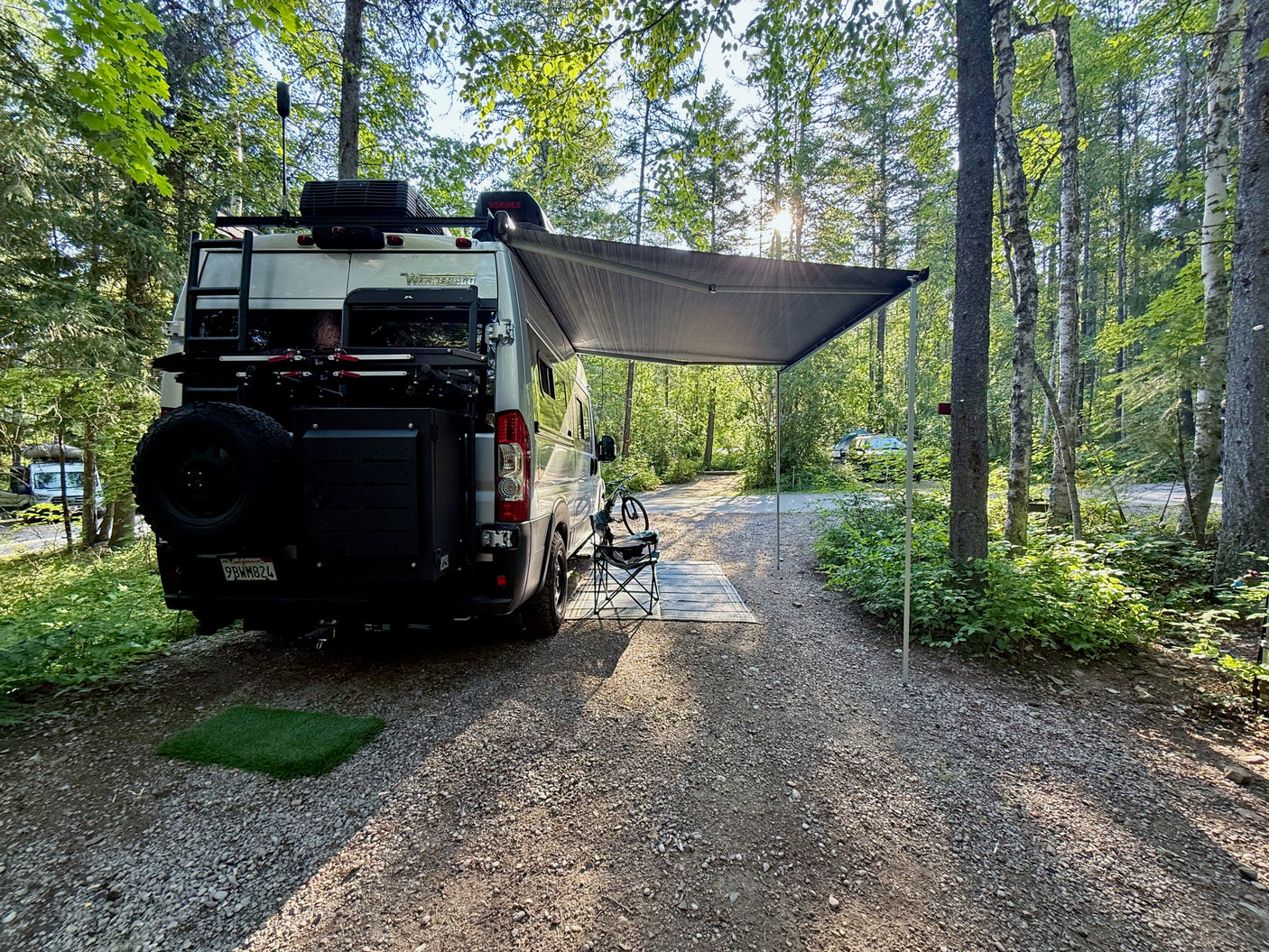 Scenic RV rental campsite view: Winnebago camper van with awning, bike, and chair in a sunny forest setting.