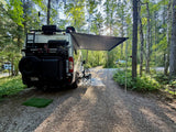 Scenic RV rental campsite view: Winnebago camper van with awning, bike, and chair in a sunny forest setting.