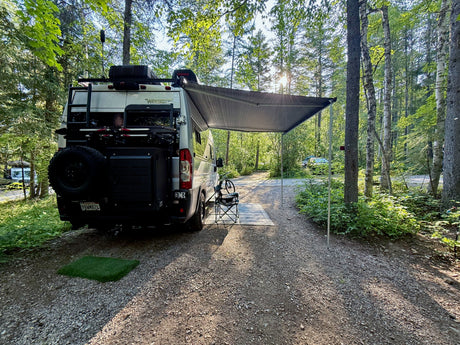 Scenic RV rental campsite view: Winnebago camper van with awning, bike, and chair in a sunny forest setting.