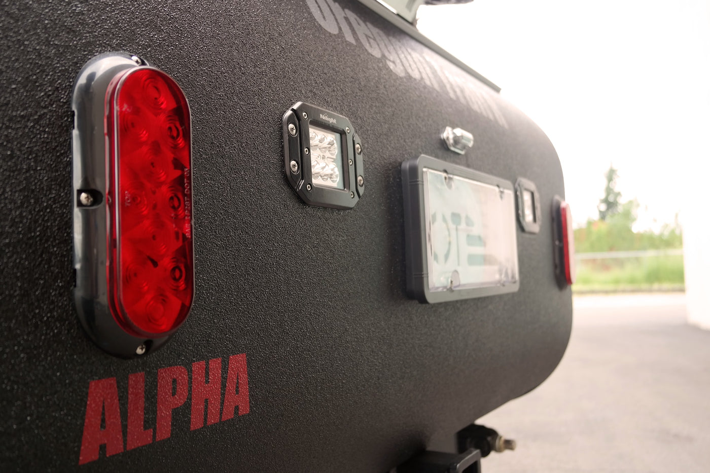 Rear of a rugged RV rental, showing a red taillight, Nilight LED, and license plate area on a black textured surface. ALPHA branding visible. — photo 3