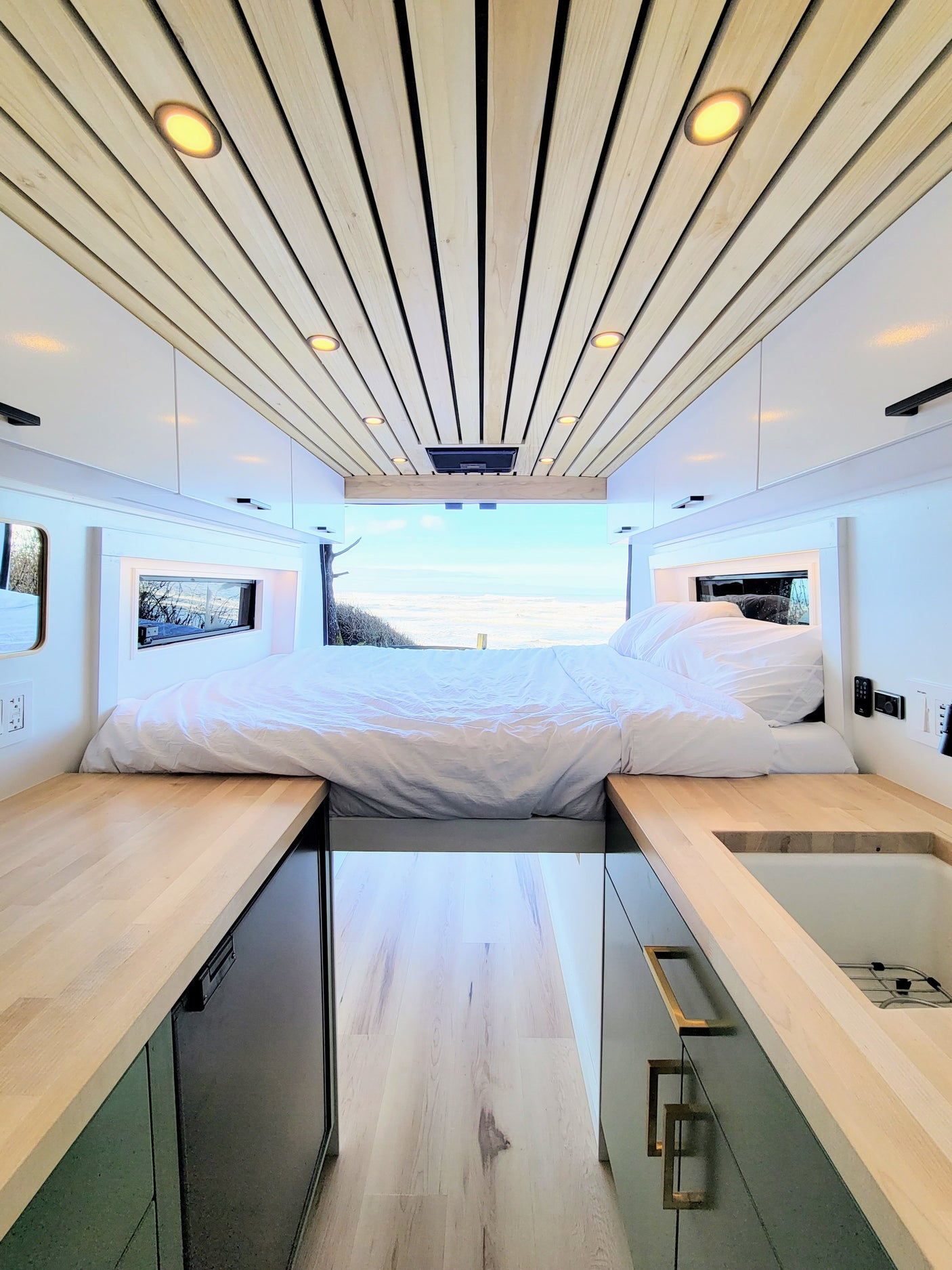 Spacious RV rental interior featuring a wood-slat ceiling, white bed, light counters, & stunning ocean view.