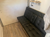 Cozy RV rental interior featuring a black futon, charging shelf, and wood-look flooring.