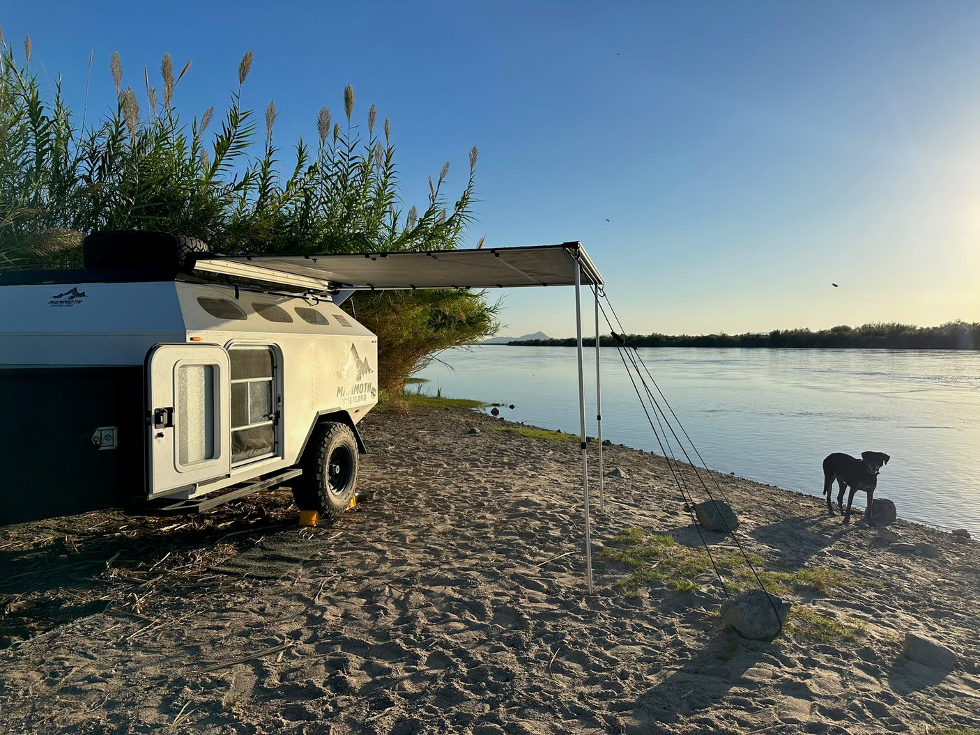 An RV rental, an overland trailer parked on a sandy riverbank with its awning extended, and a black dog by the water.
