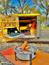 Relaxing RV rental setup: Open camper with cozy bed & outdoor kitchen. A lit fire pit sits on an orange mat.